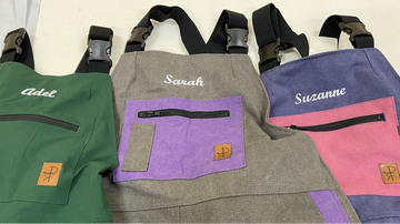 Three personalized bib overalls laid flat on a light background, each with a different body color: green, heather gray, and lavender. Each has a large front zip pocket with a contrasting pocket insert, black straps, and white embroidered names: Adel, Sarah, and Suzanne.
