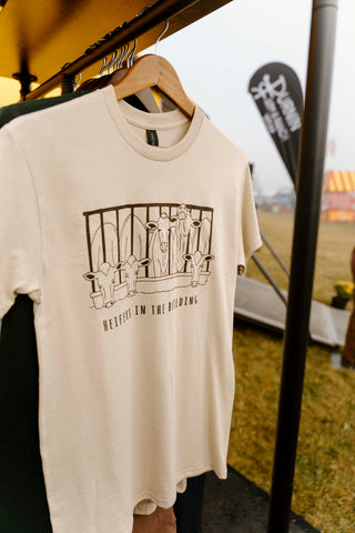 Beige t-shirt with graphic design on a hanger against an outdoor event backdrop