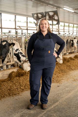 Woman in a blue overall standing in a barn with cows.