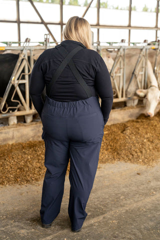 Person wearing navy overalls in a barn setting with cows and hay.